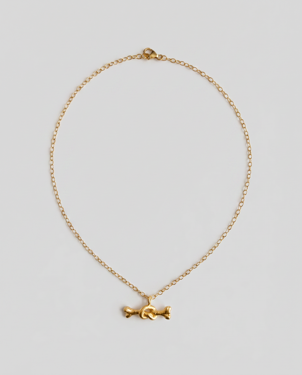 A delicate gold chain necklace featuring a small, stylized gold bone charm in the center, displayed against a solid light gray background.