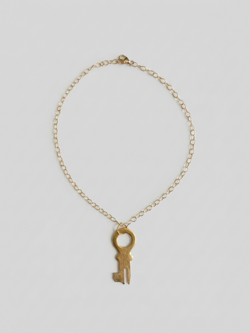 A delicate gold chain necklace featuring a vintage-style skeleton key pendant, displayed against a neutral light gray background.